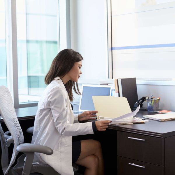 Female Doctor Wearing White Coat Reading Notes In Office